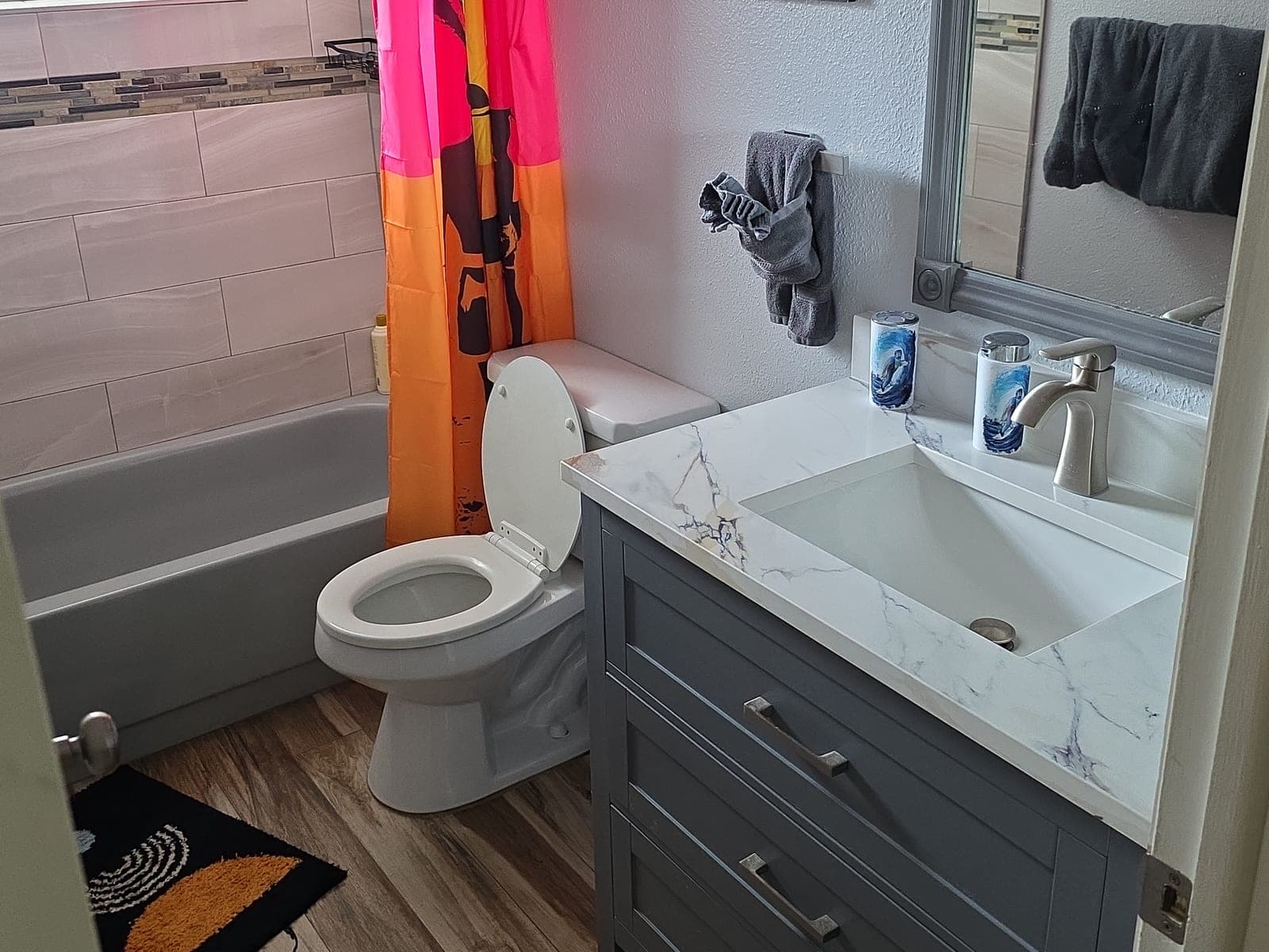 Completed bathroom with gray shaker vanity, quartz countertop, tiled tub surround, and plank flooring