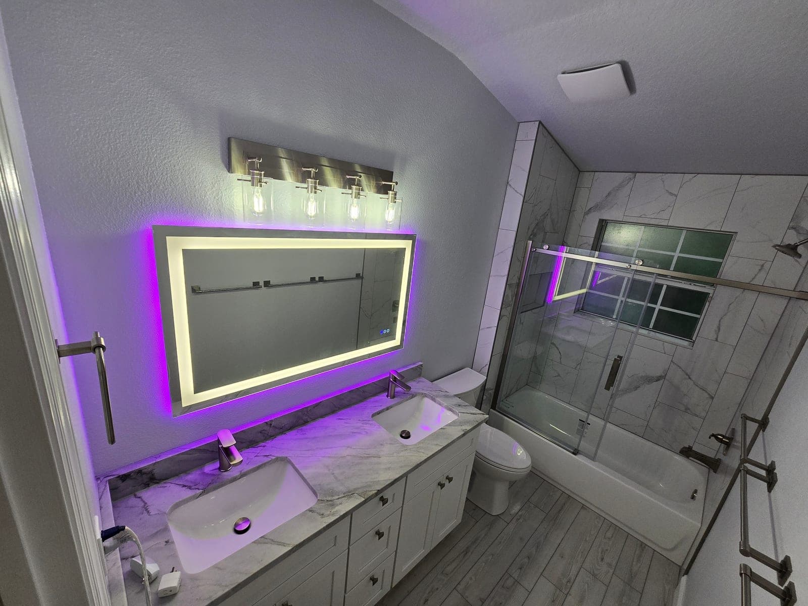 Same bathroom after remodel with marble look tile, a glass enclosed tub and shower, and a backlit LED mirror