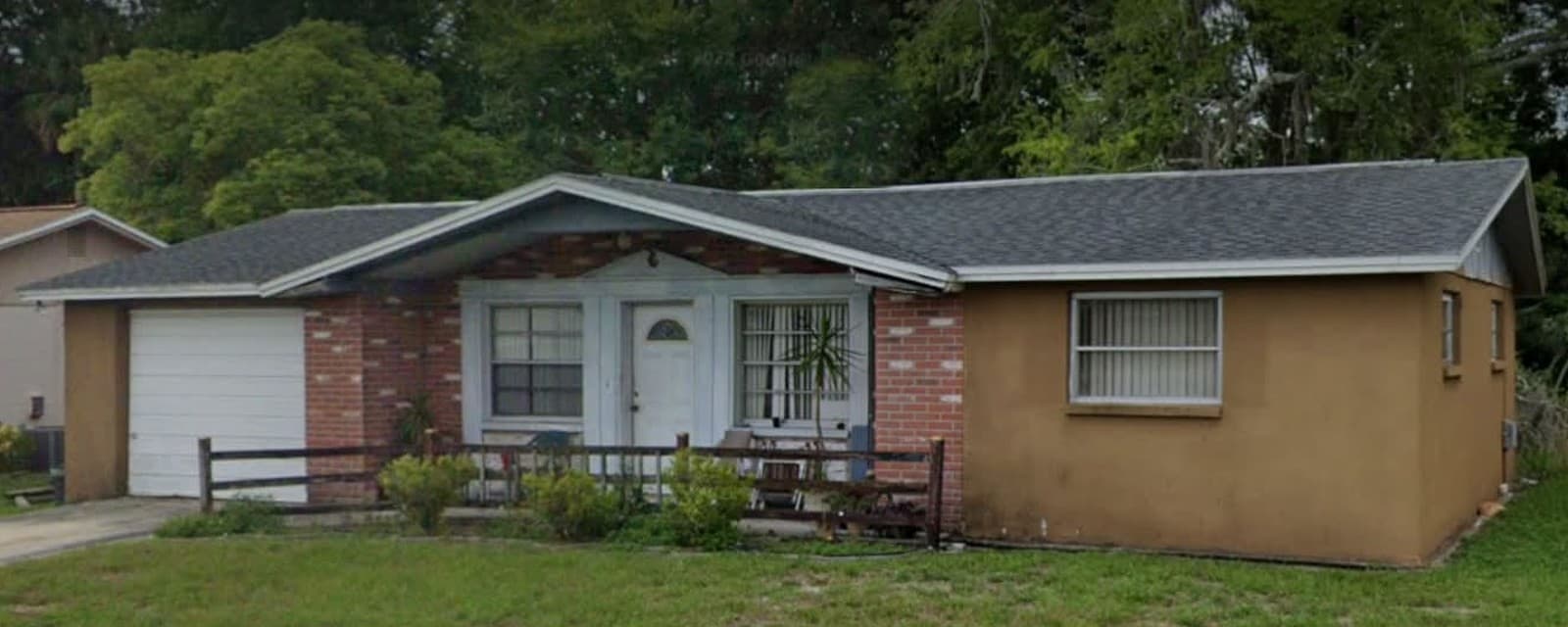 Single story home before exterior work with tan stucco, red brick around the entry, and a wood rail fence across the front porch